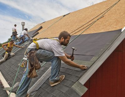 A team of men install watreproofing material and shingles to create a new roof on a rural building.